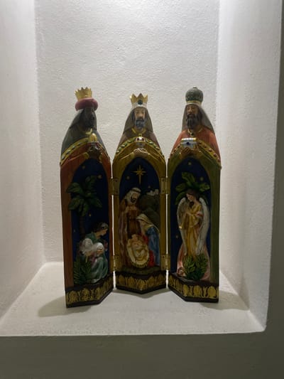 The image shows a three-panel religious triptych of the Three Kings (Three Wise Men) displayed inside a recessed niche in a wall. The triptych is made of wood and features carved figures of the kings on the outside panels. The inside panels depict scenes from the Nativity story, including an angel, the baby Jesus, and Mary and Joseph. The triptych has ornate gold detailing around the edges and is secured with hinges.
The wall and niche are painted white, and the lighting is somewhat dim, suggesting an indoor setting. The image was taken in Oaxaca City, Mexico.