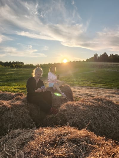 This photograph captures two women relaxing on hay bales in a rural landscape at sunset in Стеклянный, Russia. The low sun, positioned towards the right of the frame, casts a warm, golden glow across the scene, creating a noticeable lens flare and highlighting the textures of the hay.

The woman on the left, with grey hair tied in a bun, wears a dark long-sleeved jumpsuit and red sneakers. She is seated on a large hay bale, intently looking at a light blue smartphone held in both hands. Her posture suggests a moment of quiet absorption.

Beside her, slightly to the right, a second woman with blonde hair is also seated on a hay bale. She is dressed in a pink jacket and light-colored shorts, and holds a dark smartphone in her right hand, similarly focused on its screen. A white cup is visible in her left hand.

The setting is an expansive green field, crisscrossed by a dirt path. A dense line of trees forms the horizon in the background, contrasting with the soft, wispy clouds scattered across the blue sky overhead. The hay bales, golden brown from the sunlight, dominate the foreground, providing a rustic and natural seating area. The overall mood is one of tranquil leisure, as the two individuals engage with their personal devices amidst the beauty of the evening outdoors. No visible text is present in the image.