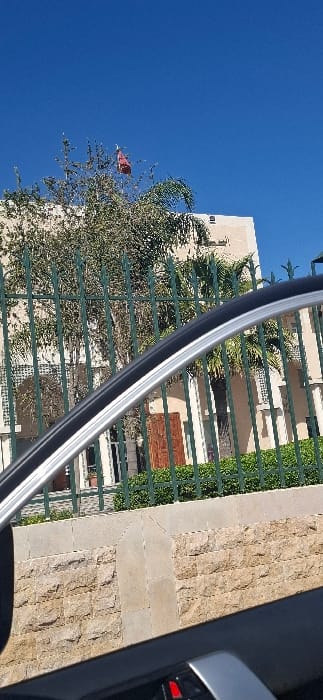 The image displays an outdoor scene in Berrechid, Morocco, viewed from within a vehicle. The foreground is defined by the silver and black trim of a car's side window or door frame, with a small red element visible in the lower right. A tall, dark green metal fence with vertical bars and pointed finials extends across the midground. Beneath the fence, a retaining wall constructed of light brown rough-hewn stone blocks with a grey concrete coping is visible. In the background, a multi-story, light-colored building, likely an institutional or official structure, is partially obscured by dense vegetation. This vegetation includes large deciduous trees and palm trees, with green shrubbery lining the base of the fence. A red flag is discernibly positioned atop the tallest deciduous tree, set against a clear, deep blue sky. No individuals are visible.