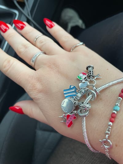 A light-skinned left hand, displaying five fingers, is prominently featured in the foreground. The fingernails are painted a glossy red and have a slightly pointed, almond shape. Several silver-toned rings adorn the fingers: a simple band on the index finger, a double-banded ring with embedded clear stones on the middle finger, and a simple band on the ring finger. A silver-toned charm bracelet is worn on the wrist, featuring various charms: a ribbed lighthouse, an airplane, a rectangular house charm colored with green, white, and red stripes (resembling the Italian flag), a blue and white striped shirt, a small heart, a skateboard, a circular disk engraved with "life force," and a pink map-shaped charm with white lettering spelling "PERU." Adjacent to the charm bracelet, a second bracelet is visible, composed primarily of small red beads, interspersed with a few silver-toned elements and light blue/turquoise rectangular beads. The background is dark and blurred, indicating the interior of a vehicle, consistent with a car's dashboard or passenger seat. The scene is located in Ulldecona, Spain.