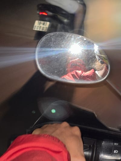 The image is a first-person perspective shot from a motorcycle or scooter in Mai Chau, Vietnam. The rider's hand, wearing a red jacket, is visible in the foreground on the right, gripping a handlebar. A rear view mirror is prominently displayed in the center, reflecting the rider, who is wearing a red jacket, sunglasses, and a helmet with gold trim. The license plate of another vehicle, which reads "18-K1 006 03", is visible on the back of a scooter in the background to the left, along with its red taillight. The scene appears to be on a street at night, indicated by the reflections and light flares, typical of urban or semi-urban environments in Mai Chau.