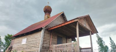 The image shows a wooden Orthodox church in Novy Izborsk, Russia. The church has a red metal roof and a wooden dome topped with a cross. The walls are made of logs. There is a covered porch with a wooden railing in front of the church. Trees are visible on either side of the church. The sky is cloudy.
