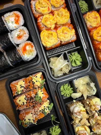 The image shows four black plastic containers of sushi. The containers are arranged on a light brown surface. The sushi appears to be of various types and fillings, including rolls topped with a yellow sauce, rolls with salmon, rolls with tobiko, and rolls wrapped in nori. Each container also includes portions of wasabi and pickled ginger. There is no indication of Tomilino, Russia or any people, actions, or interactions in the image.