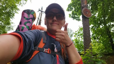 The image is a first-person perspective selfie taken outdoors, likely in a wooded or park-like setting within the city of Orekhovo, Russia. A woman, wearing a baseball cap and sunglasses, is giving a thumbs-up gesture. She is dressed in a dark blue t-shirt with red trim and is wearing a backpack with orange straps. A colorful, camouflage-patterned sleeping bag or mat is visible attached to the backpack. The background features lush green trees, a tall tree with a circular, reddish-brown object attached to its trunk, and a utility pole with electrical wires. The overall atmosphere suggests an outdoor activity or hike.