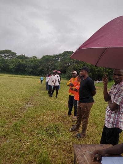 A group of people stand in a line in a grassy field in Morogoro, Tanzania, located at latitude: -6.835, longitude: 37.653. The people are standing in a line, and one man is holding a large red umbrella. There are several other people standing behind the man with the umbrella, and a couple of people are walking toward the group in the distance. The ground appears to be wet and muddy. The sky is cloudy and overcast. The scene is in a rural area, with a line of trees in the background. The people in the line appear to be registering for a race.