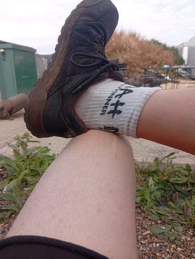 A close-up, eye-level shot shows a person's right leg, from mid-thigh downwards, resting on their left leg. The right leg is covered by a white sock with black text that reads "HI-TEC". A dark brown hiking boot is placed on top of the socked right leg, with the sole pressing into the sock. The boot is dirty, indicating outdoor use. The person is wearing dark clothing on their lower left leg, partially visible at the bottom of the frame. The background is outdoors, with a gravel path, green plants, and dry, brown vegetation. In the distance, a green utility box and some metal structures are visible under an overcast sky. The setting appears to be a semi-urban or rural area in West Suffolk, United Kingdom, possibly a garden or a yard.