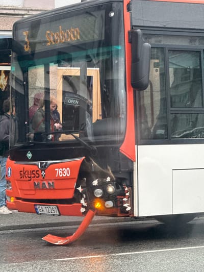 The image shows a bus that has been involved in an accident. The bus is red and white, and appears to be a public transport vehicle. The front of the bus is severely damaged, with the bumper bent outwards and broken. There are also visible damages to the headlights. The bus is on a wet, grey road, suggesting it has recently rained or is raining.
Inside the bus, several people are visible, appearing to be passengers. One passenger in the foreground is wearing a green jacket and has blonde hair, looking at their phone. Another passenger, also with blonde hair, is visible behind them. There is a sign on the bus door that says "OPEN".
The bus displays the number "7630" and the text "SKyss MAN". A license plate reads "GA 12297". An electronic display on the front of the bus shows orange text, which is difficult to fully decipher but appears to be a destination or route.
The scene is set in Bergen, Norway, as indicated by the location context. The overall mood seems somber due to the accident. The lighting suggests it is daytime, but the weather conditions are overcast or wet, contributing to a muted color palette.