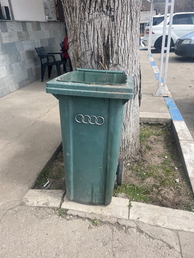 An outdoor scene in Krivodol, Bulgaria, depicts a concrete sidewalk bordering a street and a building. A dark green, wheeled waste bin, approximately one meter in height, stands prominently on the sidewalk, positioned directly against the base of a large tree with deeply furrowed bark. The waste bin's front is adorned with a silver Audi four-ring emblem, and its lid is slightly ajar.

To the left and behind the tree, a building facade is visible, constructed with rectangular grey tiles. Two dark-colored plastic chairs are placed against this tiled wall. A person wearing a red garment is partially visible seated near one of these chairs, largely obscured by the tree trunk.

To the right of the tree, a paved street or parking area is delineated by a concrete curb painted with blue and white stripes. A white SUV is parked, facing away from the viewer, and the front section of a grey vehicle is visible parked directly in front of the SUV. White metal support poles extend upwards near the curb line. The ground around the tree and between the sidewalk and curb consists of exposed soil with sparse green weeds and minor debris. The concrete sidewalk itself shows signs of cracks and wear. The background beyond the vehicles reveals a distant hilly landscape and some structures under natural lighting conditions.