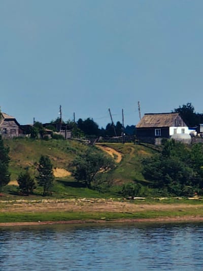 The image shows a pastoral scene with a village nestled on a grassy hill overlooking a body of water. The sky is clear and blue. On the hill, there are several wooden houses, some appearing weathered and old, with visible roofs and windows. A dirt path winds its way up the hill. Green trees and foliage are scattered throughout the landscape, with a denser cluster of trees on the right side of the hill. The foreground features the tranquil blue water. The scene evokes a sense of rural tranquility.