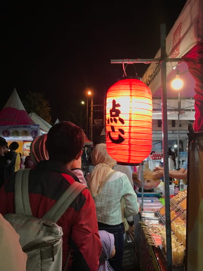 A night market stall is illuminated by a single red paper lantern hanging above a food vendor in Kecamatan Batu, Indonesia, at the coordinates -7.867, 112.514. The lantern has black Chinese characters on it. A person is wearing a red jacket, a gray backpack, and a white hat is standing in line at the stall. Another person wearing a white jacket and a beige headscarf is standing next to them. The stall has various food items, including skewers of meat, deep-fried food, and other various snacks. The scene is crowded with people, and the light from the lantern illuminates the surrounding area.