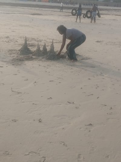 The image depicts a daytime scene on a sandy beach in Shanzu ward, Kenya. In the central foreground, an individual with dark, curly hair, dressed in a light-colored short-sleeved top and dark trousers, is crouched down, actively shaping a sand structure with their hands. This structure consists of five distinct, conical sand mounds arranged in a row. The individual is barefoot. In the mid-background, several other people are present on the beach; one figure is walking to the left, while two figures further right are carrying large, dark, circular objects resembling inner tubes or tires. The sandy surface is light-colored, showing numerous footprints and scattered small pieces of organic debris. The scene captures beach recreation.