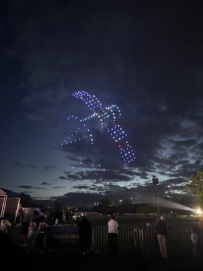 The image shows a drone light show forming the shape of an osprey, with multicolored lights illuminating the night sky in South Norfolk, United Kingdom. Below, a crowd of people stands behind metal barriers, observing the spectacle. In the background, there's a streetlight and cloudy sky. Stage equipment is partially visible on the right side of the image. The image is taken at night, with the lights of the drone show standing out against the dark sky.