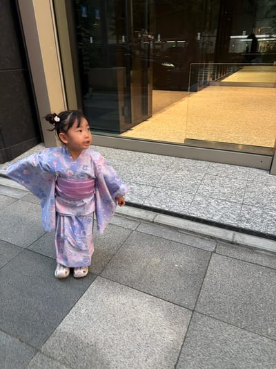 A young East Asian girl in a light purple and pink yukata with a floral and shell pattern stands on a paved sidewalk. She has her hair in two small buns with a white clip. Her hands are outstretched to her sides. The sidewalk is made of grey granite tiles with a darker grey border. Behind her, there is a large glass-fronted building with a reflective surface showing a dimly lit interior. The building has a light-colored floor visible through the glass. The overall lighting suggests it is either evening or a heavily overcast day. The scene appears to be in an urban environment, likely in the Chuo City area of Japan, given the architectural style and pedestrian area.