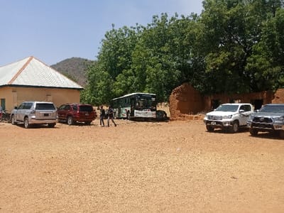 The image shows a scene in Zum, Nigeria, located at coordinates 10.116 latitude and 13.046 longitude. Four vehicles are parked on a dirt lot in front of a building with a corrugated metal roof. The building has a yellow stucco exterior. There is a large tree in the foreground, partially obscuring the view of the lot. A large, white bus is parked in the middle of the lot, with its front facing the camera. A red SUV is parked to the left of the bus and a silver pickup truck is parked on the right of the bus. Behind the bus, there are two people in the distance, walking away from the camera, and another person is walking toward the camera. There are also remnants of a brick building in the background, to the right of the vehicles. The ground is dry and dusty. The sky is blue and clear.