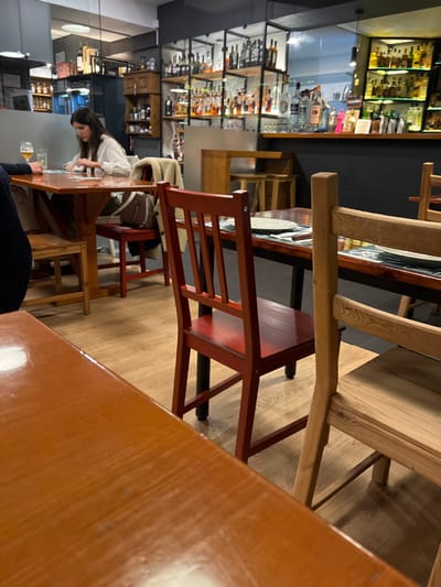 An indoor shot captures a restaurant or bar interior, focusing on wooden furniture. Two wooden tables, one partially obscured by the other, dominate the foreground. The central focus is a red wooden chair with a slated back. In the background, a woman in a white top is seated at a table, near what appears to be a partially visible man in a dark jacket. Shelves filled with bottles, possibly alcohol, line the wall, suggesting the venue is a bar or restaurant. The scene is well-lit with artificial lighting.