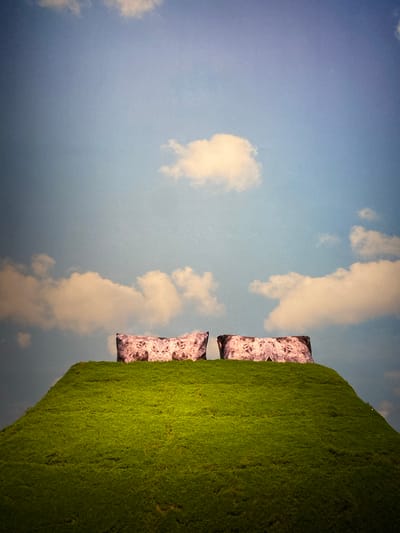 Two abstract, patterned rectangular objects, possibly pillows or cushions, are placed side-by-side on top of a vibrant green, conical mound. The mound appears to be covered in a dense, textured material resembling moss or short grass. The background is a bright blue sky dotted with several fluffy, white clouds. The scene is shot from a low angle, emphasizing the height of the mound and the expanse of the sky. The overall impression is one of surreal, minimalist landscape art. The image was taken in Jinniu District, China, but no specific identifiable landmarks within the city are visible.