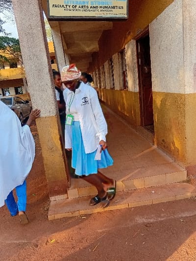 A tilted shot shows a sign for the Faculty of Arts and Humanities at Kogi State University, Anyigba. People are walking near the entrance to the building. The architecture appears to be made of stone. The outdoor steps show wear and tear. One person is wearing a Chanel-branded shirt and carrying blue papers. A second person is wearing a headwrap. The outside ground is made up of reddish soil.