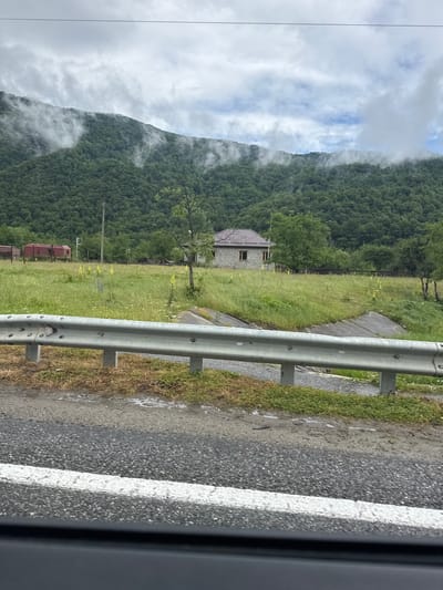 The image shows a rural landscape in Ново-Тебердинское сельское поселение, Russia, on a cloudy day. In the foreground, a metal guardrail is visible, separating the road from a grassy field. The field is dotted with yellow wildflowers and a few trees. In the middle ground, a small, light-colored house with a brown roof is situated. Behind the house, a dense, green forest covers the side of a mountain. Wisps of fog cling to the upper slopes of the mountain. To the left of the house, a red train car is visible. A wooden utility pole stands in the field. The road in the foreground has a white line marking.