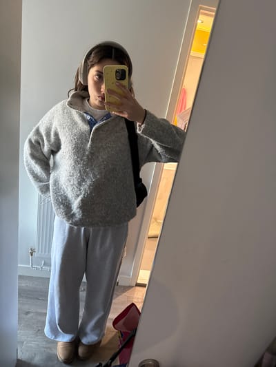 A mirror selfie captures a person, likely a young woman, standing indoors. The subject is wearing a light grey, fuzzy cardigan or jacket over a light-colored top with a visible collar, light grey loose-fitting trousers or sweatpants, and tan Ugg-style boots. Light-colored over-ear headphones are worn on her head. She holds a yellow smartphone with a studded case, obscuring most of her face except for her right eye and part of her mouth. Her fingernails are painted a light, possibly pink or white, color.

The background, reflected in the mirror, depicts a modern interior space. A white wall runs along the upper and left portions of the reflection. In the upper-middle, a horizontal rectangular recess or opening, possibly a shelf or cabinet, is brightly illuminated with a warm yellow light. Inside this recess, neatly folded pink and white items, appearing to be towels or clothing, are visible. To the right of the recess, a white wall with a silver door handle can be seen. The lower part of the reflection reveals light grey wooden or laminate flooring. A white radiator with exposed pipes is visible on the lower left side of the floor, and part of a red bag or basket is seen on the lower right, near the person's feet. The activity is a casual mirror selfie, likely taken in a home setting in Hull, United Kingdom. No visible text is present.