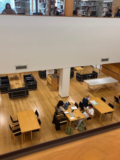 The image depicts a multi-level library interior within the Faculty of Arts and Humanities (Faculdade de Letras) building of the University of Porto, located in Lordelo do Ouro e Massarelos, Portugal.
The scene, viewed from an elevated perspective, features light-colored wooden flooring throughout. The upper level, delineated by a white wall and a wooden railing, shows a row of six individuals, predominantly women, seated at individual desks facing into the space. Each is engaged with a laptop computer. Behind them, bookshelves line the walls, visible through open sections.
The lower level is an expansive study and lounge area. In the foreground-midground, two large rectangular wooden tables with black-framed chairs are present. One table is empty with six chairs. The other table accommodates three individuals: a woman with dark curly hair using a laptop and tablet on the left, a man in a light shirt reading a book or document on the right, and a woman with blonde hair using a laptop in the background. Items on this table include laptops, notebooks, pens, a water bottle, and a green backpack.
Further into the lower level, two distinct lounge areas are arranged with black leather armchairs and sofas featuring chrome frames, each accompanied by a small wooden coffee table. A tall, multi-tiered wooden display unit containing various small objects stands near a central white support pillar. To the far right, a large rectangular table with a white top and a unique wooden base is visible. The far right wall is paneled with wood. White walls and ceilings complete the environment, with air vents visible on the white ledges. All individuals are focused on their respective tasks, indicating independent work or study.