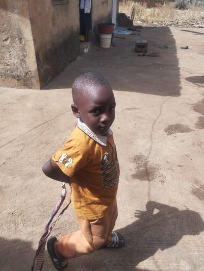An outdoor scene in Unguwan Rimi, Nigeria, depicts a young child kneeling on a concrete or packed earth surface, directly facing the viewer. The child, with dark skin and closely cropped hair, wears an orange-gold short-sleeved collared shirt with various emblems and text, including "UNBLOCKED" on the collar and a numeric "7" on the right sleeve. Beige trousers and black and white striped slide sandals complete the attire. A patterned, multi-colored strap or fabric piece is visible extending from behind the child's left side. To the left, a weathered light brown building with a shadowed doorway reveals the lower legs of an individual wearing yellow sandals. Near the doorway, a red basin sits atop a white bucket. The ground surface varies, with patches of moisture or staining. In the background to the right, scattered debris, a wooden block, and a natural fiber broom are visible on the ground. Beyond this, an unkempt area with sparse, dry vegetation extends to the horizon. Strong daylight casts distinct shadows, including that of the child, on the ground to the right.