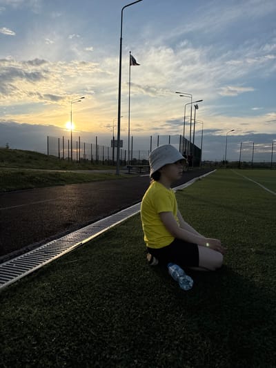 The image shows a young person in a yellow t-shirt and black shorts sitting on green grass near a running track. They are wearing a grey bucket hat and have their legs bent with their hands clasped together. A plastic water bottle is placed on the grass next to them. The background features a soccer field with white lines, a fence, and several lampposts. The sky is blue with wispy clouds, and the sun is setting, casting a warm glow over the scene. The overall impression is one of relaxation or a break during physical activity. The location appears to be an outdoor sports facility in Gornoe, Russia, given the context.