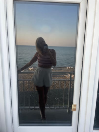 The image shows a reflection in a glass door of a woman taking a selfie. She is standing on a balcony overlooking a beach and the sea. The sky is clear with the sun setting. The woman is wearing a striped crop top and white shorts. On the beach, there are several empty lounge chairs. The scene is captured from a slightly elevated perspective, suggesting the balcony is on an upper floor. The location is identified as Giachrypsh, Abkhazia.
