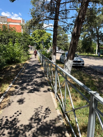 A person is walking on a paved walkway bordered by a metal fence and lush greenery. In the background, a white BMW is parked on the side of a road, with other cars and buildings visible further down. The sky is clear and blue with scattered clouds, and sunlight casts dappled shadows from the trees onto the walkway. The scene appears to be a public area in Timashevsk, Russia, likely a park or a quiet street.