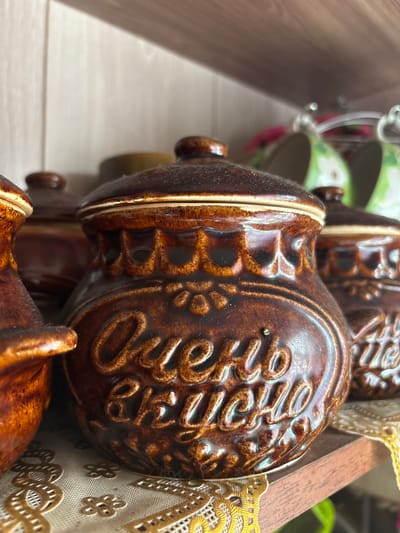 The image shows a close-up view of a set of brown ceramic pots with lids, arranged on a wooden shelf. The pots have an embossed pattern on their surface and appear to be decorated with a floral motif. The front-most pot has Cyrillic text embossed on it, reading "Очень вкусно", which translates to "Very tasty". The pots are placed on a doily or tablecloth with a floral pattern. In the background, other ceramic items and possibly mugs are visible. The lighting suggests an indoor setting, likely a kitchen or dining area. The scene is static and depicts decorative kitchenware. There is no indication of the city of Ветлужский, Russia.