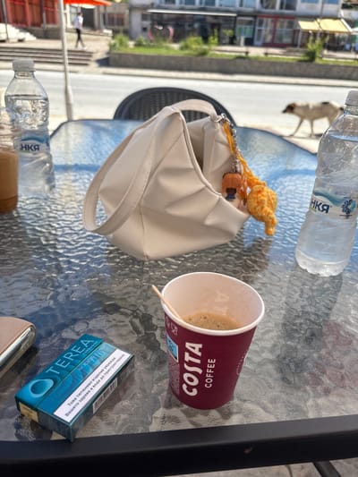 An outdoor table at a cafe in Garmen, Bulgaria, is set with a coffee cup, a pack of Terea Turquoise for IQOS Iluma cigarettes, a white purse with a keychain, and two bottles of water. A person is walking in the blurred background, and a dog is walking on the street. The table has a glass top with a textured pattern, and it is positioned on a patio overlooking a street with buildings and trees. The lighting suggests it is daytime.