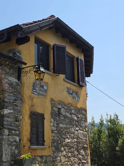 The image shows a close-up, exterior view of an aged, yellow-plastered building with stone accents in Orta San Giulio, Italy. The building features dark wooden shutters on its windows. A wrought-iron lantern hangs on the wall near a window. The lower portion of the building is constructed with rough stone masonry. A small sign with the number "17" and Italian text is affixed to the stone wall. In the foreground, a planter box with pink and red flowers is visible. The background shows lush green foliage and a clear blue sky. The overall impression is of a rustic, historic structure in a Mediterranean setting.