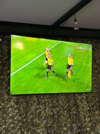 The image shows a close-up of a television screen displaying a soccer match. The screen is mounted on a wall covered with artificial greenery. The television shows three figures in yellow and black referee uniforms on a bright green soccer field. Two of the figures are walking together, while the third, in the foreground, is holding a soccer ball and a yellow and red checkered flag. A timer in the bottom right corner of the screen reads "20:06." In the top right corner of the screen, there is text that appears to be in Cyrillic script, with the word "НА ЖИВО" meaning "LIVE". The television is positioned in what appears to be an outdoor seating area, indicated by the ceiling structure and exposed light bulbs visible in the background. The overall setting suggests a sports bar or restaurant with an outdoor viewing area. The image is taken from a low angle, looking up at the screen. The location is identified as Ravno pole, Bulgaria.