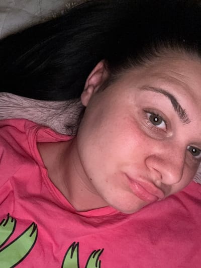 A close-up, overhead, indoor shot shows a woman with dark hair, fair skin, and green eyes. She is lying down, with her head resting on a textured, light-colored surface, likely bedding. Her eyes are open and looking upwards. She is wearing a bright pink t-shirt with a graphic design of green cartoon-like figures on the front. Her facial expression is neutral, with her lips slightly pursed. The lighting in the image is dim, casting soft shadows on her face and hair. The background is predominantly dark due to her hair. This image was taken in Rogosh, Bulgaria.