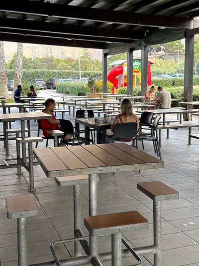 This image depicts an outdoor dining area, likely at a McDonald's restaurant in Carini, Italy, given the presence of a McDonald's PlayLand structure in the background. The area is covered by a dark wooden pergola. Several tables and chairs, typical of fast-food establishments, are arranged on a tiled floor.
In the mid-ground, two women are seated at a table. One woman, wearing a red top, is facing away from the camera, and the other, wearing a dark top, is also facing away. Further back, a man and a woman are seated at another table. The man is wearing a light-colored t-shirt, and the woman is wearing a white top. Both appear to be engaged in eating or drinking.
In the left mid-ground, a man in a blue shirt is seated at a table, facing a woman in a white top. They are also seated under the pergola.
The background reveals a playground area with a red slide and colorful structures, identified as "PlayLand." Beyond the playground, there is a line of cars parked in what appears to be a parking lot, bordered by green hedges and trees. In the far distance, a mountainous landscape is visible under a hazy sky. The overall atmosphere suggests a casual dining setting, possibly during daylight hours.