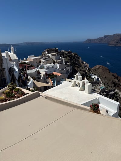 The image shows a scenic view of the village of Oia in Santorini, Greece, overlooking the Aegean Sea. The foreground is dominated by the beige canvas of a rooftop, with a potted plant containing red flowers and a white chair on the right. In the middle ground, a cluster of whitewashed buildings with blue accents cascades down a cliffside. Several people are visible walking along narrow pathways between the buildings. A prominent white bell tower with three arches stands out on the left side of the middle ground. The background features the deep blue sea, dotted with a few boats, and distant islands and cliffs under a clear blue sky. The architecture is characteristic of the Cycladic islands, with cubic houses and churches built into the volcanic rock. The overall impression is one of a sun-drenched, picturesque Mediterranean village.