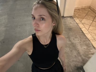 A young, fair-skinned woman with blonde hair is standing indoors, taking a selfie. She is wearing a black tank top and a black skirt, along with a gold necklace and a delicate bracelet. Her left arm is visible, showing freckles. The background consists of a tiled floor and a neutral-colored wall, with a column to her right. The scene appears to be in a hallway or a similar indoor public space. The lighting is dim, with some reflections on her skin.