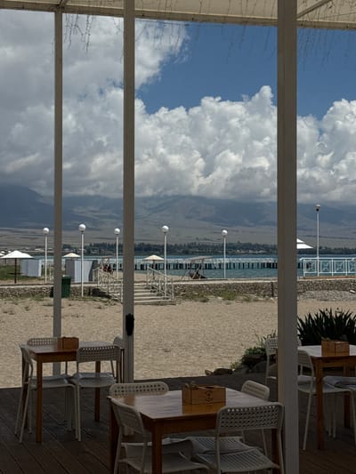 The image depicts an outdoor seating area at a beach resort in Koshkol', Kyrgyzstan. In the foreground, wooden tables with white chairs are arranged on a wooden deck. A sandy beach with scattered stones and sparse vegetation extends beyond the deck. In the midground, a paved walkway leads to a body of water, likely Issyk-Kul lake, with a pier stretching into it. Several lampposts are visible along the shoreline. In the background, rolling hills and mountains under a cloudy sky complete the landscape. There are no people clearly visible in the immediate foreground, though figures can be seen in the distance near the pier and seating areas. The overall scene suggests a tranquil, scenic resort environment.