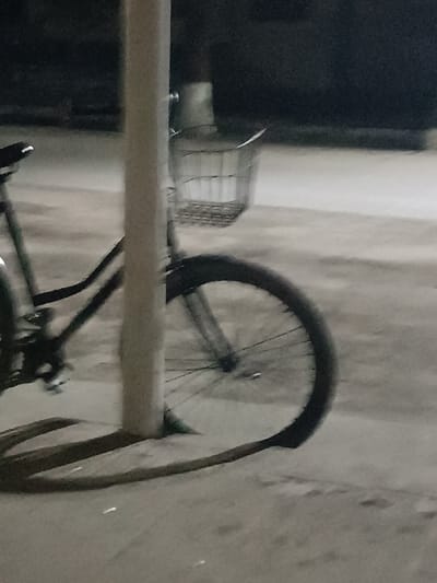 The image captures a close-up, somewhat grainy view of a bicycle parked outdoors, predominantly at night or in very dim lighting. A thick, light-colored vertical pole stands prominently in the center foreground, obscuring the middle section of the bicycle.
To the left of the pole, parts of the bicycle's dark-colored frame, a portion of the seat, and what appears to be the handlebars are visible. Further down on the left, the pedals and chainring mechanism are also discernible.
To the right of the pole, the front wheel of the bicycle is clearly in view. It features a black tire with visible spokes. Directly behind the pole and above the front wheel, a wire basket, likely attached to the handlebars or front fork, is visible. It appears empty.
The bicycle is situated on a paved surface, which looks like concrete or asphalt, exhibiting a light gray tone. A distinct crescent-shaped shadow or perhaps a light reflection is cast on the ground, extending from the base of the central pole towards the bottom right of the frame. The background is very dark and blurry, suggesting either deep shadow or the low-light conditions of nighttime. There are no other discernible objects or people in the scene. The overall impression is one of a bicycle parked and left alone in a quiet, dimly lit area.