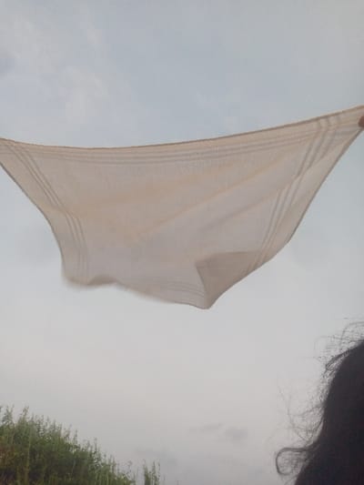 A cream-colored, slightly soiled handkerchief is held taut against a cloudy sky. The fabric shows a subtle striped pattern near its edges. Below, a glimpse of green vegetation is visible, and the edge of a person's dark hair enters the frame on the right. The overall impression is one of simplicity and slightly muted colors.