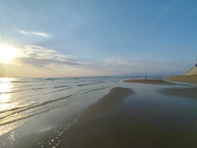 The image depicts a beach scene in Agios Stefanos, Greece, at sunset. The sun is low on the horizon, casting a bright glare and long reflections on the wet sand and shallow waves. The sky is pale blue with scattered clouds. The beach is wide and sandy with a person walking in the distance near the water's edge. A small stream or channel flows across the beach towards the sea. A cliff or elevated landform is visible at the right edge of the frame.