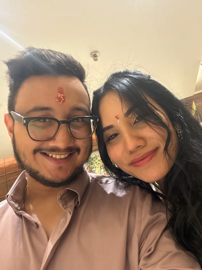 A couple poses closely together for a selfie. The woman, with long dark hair and a small dot on her forehead, smiles warmly. The man, wearing glasses and sporting a beard, also smiles broadly. Both are wearing matching light pink shirts. A few grains of rice stick to the man's forehead. The background is blurred, suggesting an indoor setting. The image is rotated, with the subjects appearing sideways.