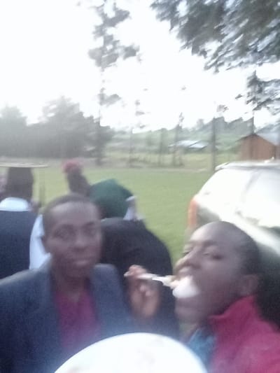 An outdoor gathering features a man in a blazer and purple shirt, and a woman eating from a spoon. Other people stand behind them. A white car is on the right, and a building in the distance. Trees and foliage are in the background, and a blurred sky is overhead. The image is slightly out of focus.