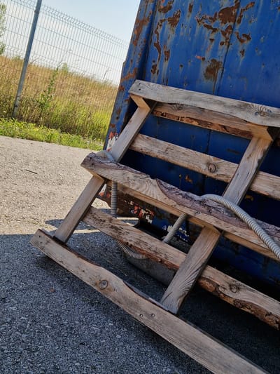 The image captures an outdoor scene with a focus on a leaning, weathered blue dumpster against a backdrop of a chain-link fence and dry vegetation. In the foreground, a crude wooden structure, possibly a makeshift support or ramp, is propped against the dumpster. This wooden assembly is constructed from rough-hewn planks, showing signs of age and wear, with some exposed bark. A ribbed, grey electrical conduit snakes across the wooden structure and disappears behind the dumpster. The ground is a gravelly surface. The overall impression is one of an industrial or utilitarian area, possibly in a rural or semi-urban setting like Bahovitsa, Bulgaria, given the fence and dry landscape. No people are visible in the frame. The lighting suggests it is daytime, with sunlight casting shadows.