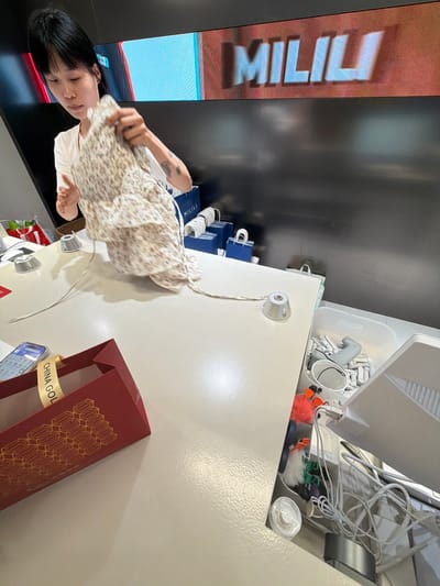 A young Asian woman with short black hair, wearing a white t-shirt, is standing behind a light-colored counter in what appears to be a retail environment. She is holding a floral-patterned fabric garment. In the background, a large digital screen displays the word "MILILU" in large, stylized letters. To the left of the woman, a red gift bag with a gold ribbon and hexagonal pattern is visible. On the right side of the counter, a clear plastic bin contains various electronic cables and small white plastic components. A smartphone is placed on the counter near the gift bag. The overall setting suggests a store or a stockroom. The specific location of Qinhuai District, China, cannot be determined from the visual information provided in the image.