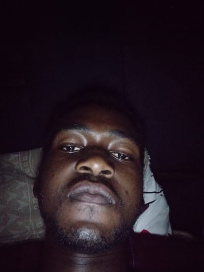 A close-up, low-light portrait captures a dark-skinned individual, likely male, appearing to be lying down and looking directly into the camera. The face, featuring dark eyes, a broad nose, and full lips, is centrally positioned in the lower half of the frame. Short, dark facial hair forms a beard and mustache around the mouth and chin. Reflections are visible on the forehead, nose, and lips, indicating a direct light source. To the left of the individual's head, a section of a light-colored, patterned pillow or bedding is visible. On the right, near the neck, a white garment with dark horizontal stripes is partially visible, layered over a darker, possibly red or maroon fabric. The background and the upper portion of the image are predominantly dark, with subtle image noise, suggesting a dimly lit indoor environment.