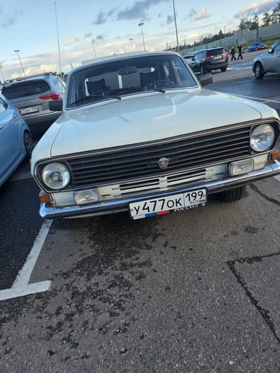 The media file shows a close-up, slightly tilted perspective of the front of a white vintage car parked in what appears to be a parking lot. The car has a prominent chrome grille, a round headlight, and a Russian license plate that reads "У477ОК 199". The license plate also features the text "RUS" and "РОССИЯ". Behind the white car, other vehicles are visible, including a silver SUV with a license plate that reads "572ВТ 250". The scene suggests an outdoor setting, possibly a parking lot in Sholokhovo, Russia, given the provided location context. The lighting indicates it might be daytime, with the sky visible in the background. The overall impression is of vehicles parked in a lot, with no specific activity or event clearly depicted beyond the act of parking.