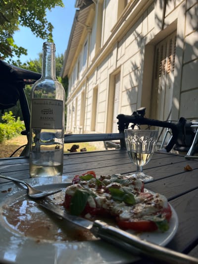 In Bièvres, France, on a wooden table outside a building, a partially consumed bottle of white wine and a glass of white wine sit next to a plate of Caprese salad. The salad consists of sliced tomatoes, fresh mozzarella, and basil, drizzled with balsamic glaze. A fork and knife are also on the plate, indicating the meal is in progress. In the background, the facade of a light-colored building is visible, with trees and foliage surrounding the area. The scene suggests a leisurely meal enjoyed outdoors.