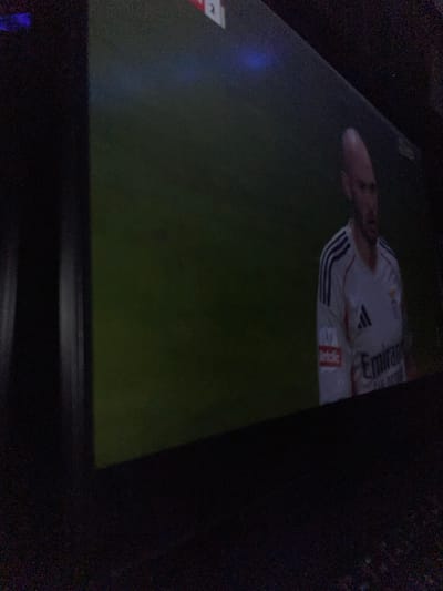 A television screen displays a soccer game with a player wearing a white Real Madrid jersey with "Emirates" printed on the front. The player's face is visible, and the soccer field is green. The surrounding room is dark, with the TV screen angled slightly upwards.