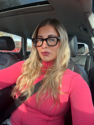 A female individual is seated in the driver's side of a vehicle, looking directly forward at the camera. She possesses long, wavy blonde hair parted on her left, dark eyebrows, and wears dark-framed rectangular eyeglasses. Her lips are coated with a dark red or burgundy lipstick. She is dressed in a long-sleeved, high-collared top or jacket of a bright coral or hot pink hue, featuring a partial zipper and visible stitching details. A black automotive seatbelt diagonally crosses her chest and shoulder. The vehicle's interior features black leather seating with headrests and a sunroof visible overhead. Direct sunlight illuminates the scene, casting shadows on the woman's face and clothing. Through the vehicle's windows, a clear blue sky is visible above a landscape with bare-branched trees. In the mid-ground, a multi-story building with prominent red architectural components and dark windows is partially visible behind the trees. The scene is located in the city of Marcq-en-Barœul, France.