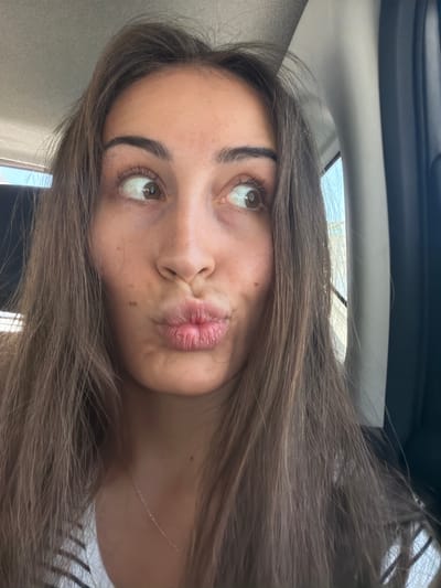 The image is a close-up, self-taken photograph of a young woman with long, brown hair. She is wearing a white and black striped top and a thin gold necklace. Her lips are pursed in a pout. Her eyes are looking to her right. The background appears to be the interior of a car, possibly in Larnaca, Cyprus, with a window and a car door visible. The lighting suggests it is daytime.